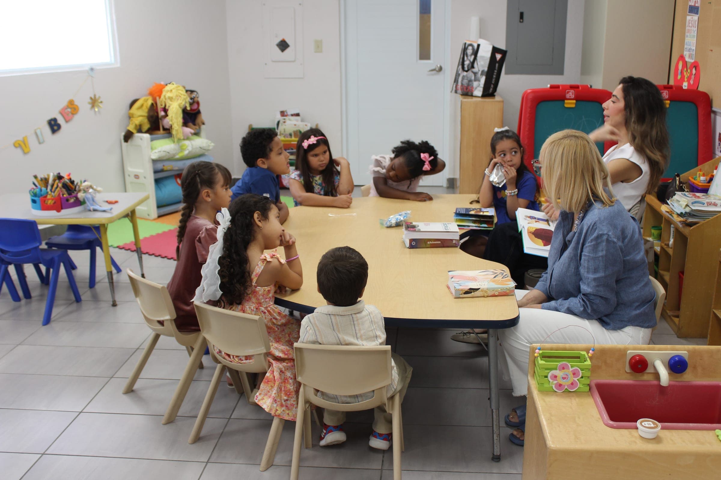Niños sentados alrededor de una mesa con sus maestras durante la clase.